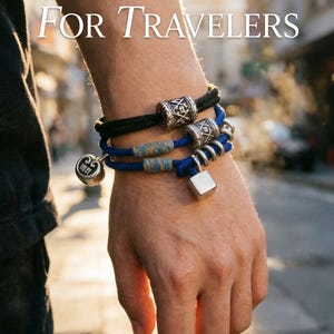 Peut inclure: Trois bracelets ornés de perles et de breloques décoratives. L'un est noir, un autre bleu avec un motif tissé, et le troisième bleu avec un charm argenté en forme de cube. Le texte "FOR TRAVELERS" est affiché en haut.