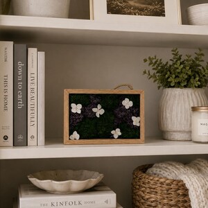 May include: A rectangular wooden frame containing a preserved moss arrangement with deep green and purple textures, accented by five small white hydrangea blossoms. The piece features a thin jute rope loop at the top for hanging display.