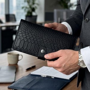 May include: A black leather wallet with a reptile skin texture is held open, revealing a dark blue leather document holder. A pen, a cup, and a notebook are on a wooden desk. The person is wearing a gray suit.