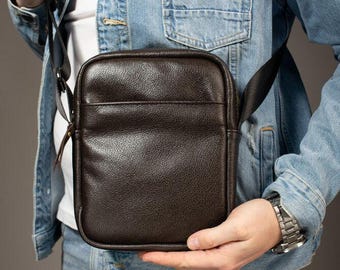 Può includere: Una borsa a tracolla rettangolare marrone scuro con una finitura testurizzata e granulata. La borsa presenta una tasca frontale con cerniera, uno scomparto principale con chiusura a zip e una tracolla regolabile in tessuto nero sui lati.