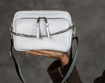 Puede incluir: Un bolso cruzado rectangular blanco con una cadena plateada y una correa estampada en blanco y negro. El bolso tiene dos cremalleras y una superficie texturizada. El bolso está sostenido por una mano.
