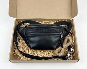 May include: A black leather fanny pack with two zippered compartments and a black adjustable strap with a plastic buckle. The bag is placed inside a brown cardboard box filled with light-colored wood shavings for protective packaging.