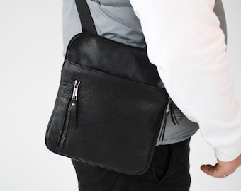 May include: A black crossbody bag with a textured surface, featuring a vertical zippered front pocket with a silver-toned metal pull, a main zippered compartment, and an adjustable black shoulder strap worn across the torso.
