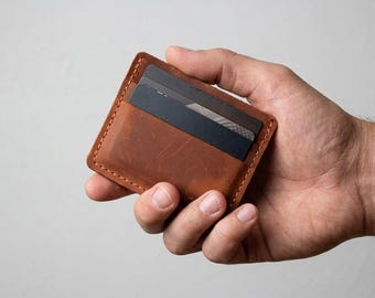 Può includere: Un porta carte rettangolare in pelle marrone con cuciture visibili lungo i bordi. Il porta carte contiene due carte grigio scuro inserite nella fessura superiore, mostrando una texture liscia e opaca sulla superficie in pelle.