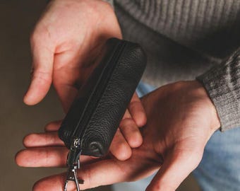 May include: A small, cylindrical black leather pouch with a textured grain finish. The pouch features a central black zipper closure running along its length and a silver-toned metal lobster clasp attached to one end by a short leather strap.