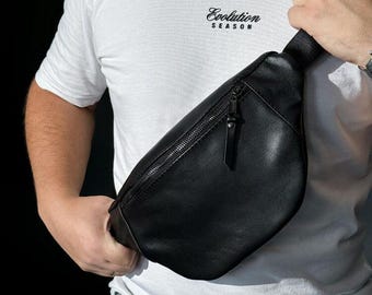 May include: A black leather-like crossbody fanny pack featuring a smooth texture, a single main compartment with a silver-toned zipper closure, and an adjustable black strap with a metallic buckle, worn across the chest over a white t-shirt.