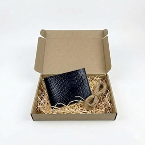 May include: A black bifold wallet with a textured snakeskin pattern placed inside an open brown cardboard box. The box is filled with light brown shredded paper filler and includes a small coil of natural jute twine resting next to the wallet.