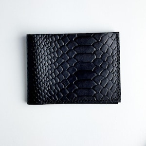 Könnte beinhalten: Schwarzes zweifach gefaltetes Portemonnaie mit einem strukturierten Schlangenhautmuster. Das Portemonnaie ist rechteckig und scheint aus Leder zu sein. Das Portemonnaie ist geschlossen und liegt auf einer weißen Oberfläche.
