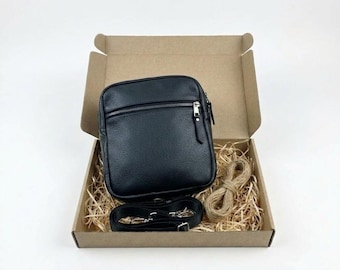 May include: A black leather crossbody bag with a zippered front pocket and a matching detachable shoulder strap. The bag is presented inside an open cardboard box filled with shredded paper filler and a coiled piece of natural jute twine.