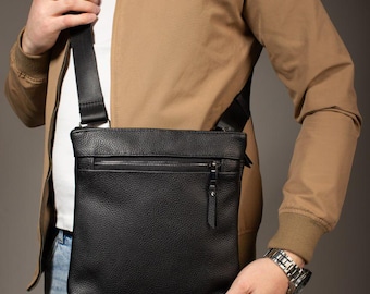 May include: A black, rectangular crossbody bag with a textured, pebbled finish. The bag features a horizontal zippered pocket on the front, a matching black adjustable shoulder strap, and a silver-toned zipper pull with a black leather tab.