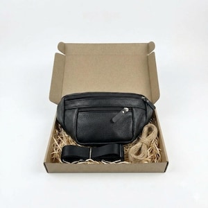 May include: A black textured fanny pack with multiple zippered compartments and a matching black adjustable strap, presented inside an open brown cardboard box filled with light-colored wood shavings for protective packaging.