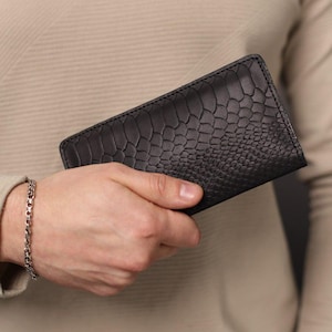 Puede incluir: Cartera negra con un patrón texturizado similar a la piel de reptil. La cartera se sostiene en una mano, mostrando su diseño. Parece estar hecha de cuero o un material similar. Se ve una pulsera plateada en la muñeca.