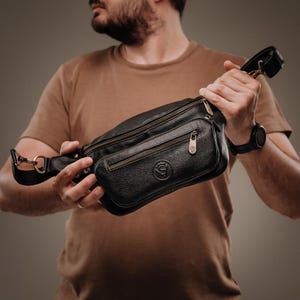 May include: A black leather fanny pack featuring a textured surface, two main zippered compartments, and a smaller front zippered pocket. The bag includes a metal logo emblem on the front, brass-toned hardware, and an adjustable strap with clips.