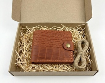 May include: A brown bi-fold wallet with a textured crocodile-skin pattern and a brass-colored snap button closure, nestled inside a cardboard gift box filled with shredded paper filler and accompanied by a small coil of natural jute twine.