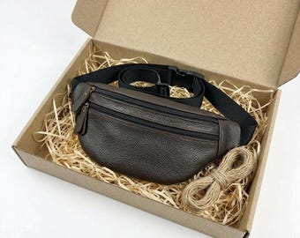 May include: A dark brown leather fanny pack with a textured surface, featuring two zippered compartments and a black adjustable nylon strap with a plastic buckle. The bag is placed inside a cardboard box filled with light brown wood shavings.