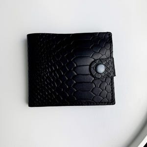 Puede incluir: Una cartera negra de dos pliegues con un patrón texturizado de piel de serpiente. La cartera rectangular tiene esquinas redondeadas y un cierre de botón a presión. El botón a presión es de color gris claro. La cartera está hecha de un material similar al cuero.