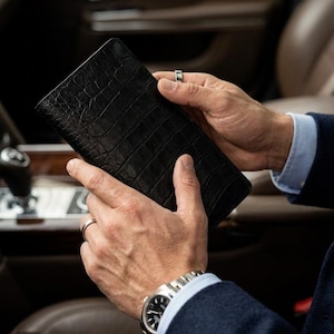 May include: A rectangular black wallet featuring a textured crocodile skin pattern. The wallet is held in a vertical orientation, showcasing its deep black color and the detailed, raised scales characteristic of the embossed leather surface.