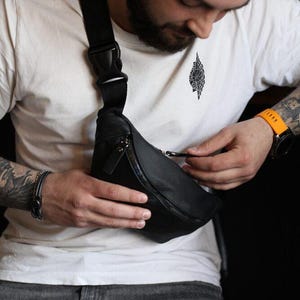 May include: A black leather fanny pack featuring a main zippered compartment with a silver-toned metal zipper pull. The bag is attached to a wide, adjustable black nylon strap with a sturdy plastic side-release buckle for secure fastening.