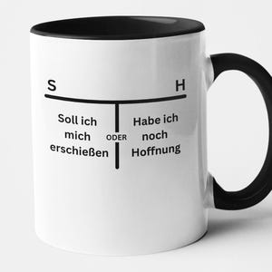 Könnte beinhalten: Weißer Keramikbecher mit schwarzem Innenraum und Henkel. Der Becher zeigt schwarzen Text auf Deutsch mit den Worten "Soll ich mich erschießen oder Habe ich noch Hoffnung" auf der Vorderseite.