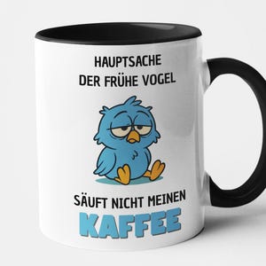 May include: White ceramic mug with a black handle and interior. The mug features a cartoon blue bird with a sleepy expression and German text that reads "HAUPTSACHE DER FRÜHE VOGEL SÄUFT NICHT MEINEN KAFFEE."