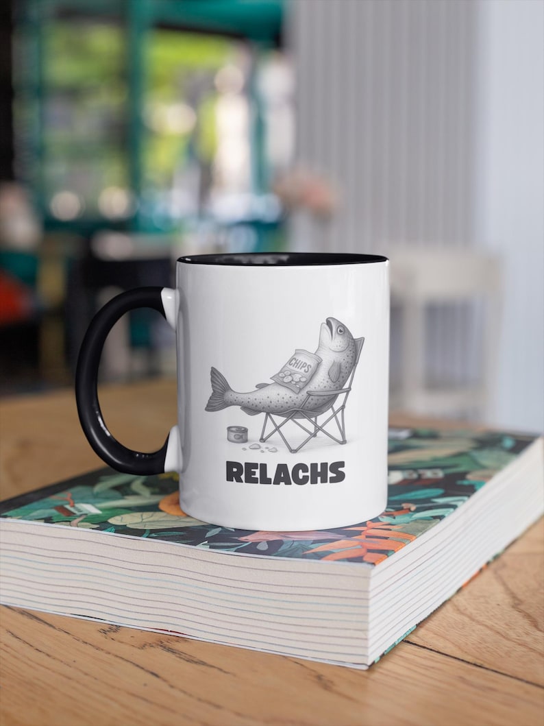 May include: White ceramic mug with a black handle and rim, featuring a humorous illustration of a fish relaxing in a chair, eating chips. The word "RELACHS" is printed below the fish. The mug is resting on a book.