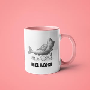 May include: White ceramic mug with a pink interior and handle. The mug features a black and white illustration of a fish relaxing in a beach chair, holding a bag of chips. The word "RELACHS" is printed below the illustration.
