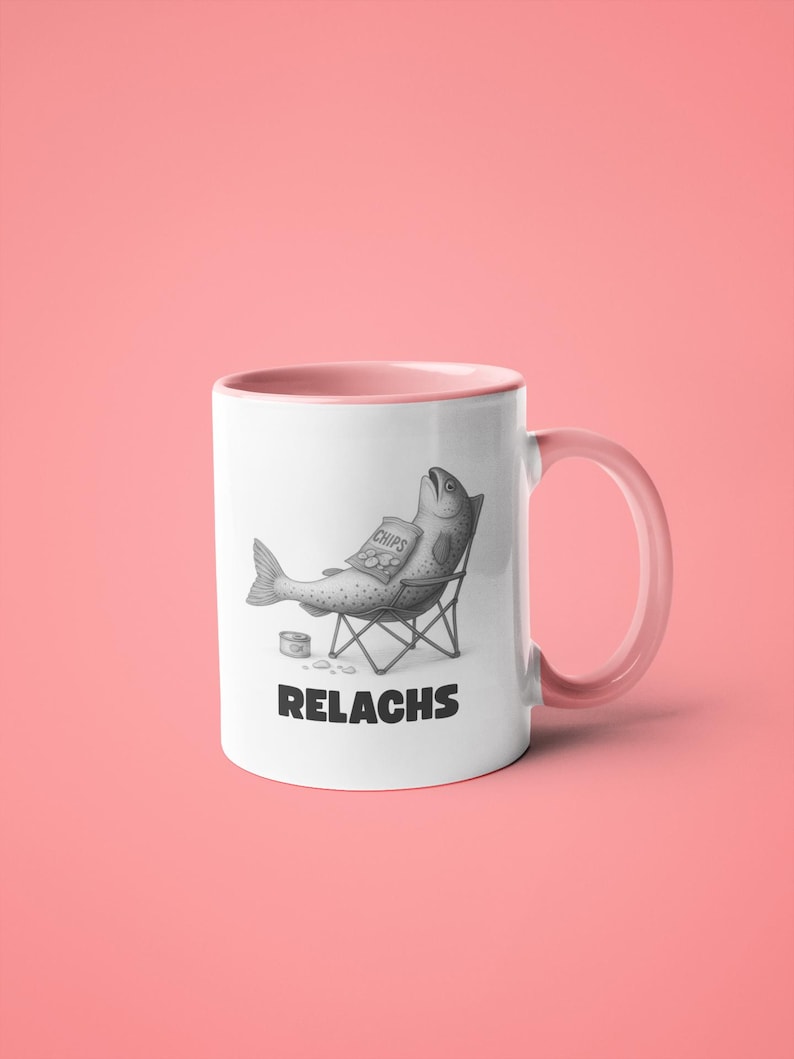 May include: White ceramic mug with a pink interior and handle. The mug features a black and white illustration of a fish relaxing in a beach chair, holding a bag of chips. The word "RELACHS" is printed below the illustration.