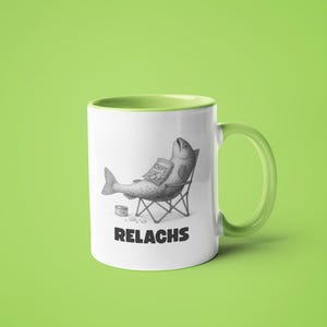 May include: White ceramic mug with a green handle and interior. The mug features a humorous illustration of a fish relaxing in a beach chair, holding a bag of chips. The word "RELACHS" is printed below the illustration.
