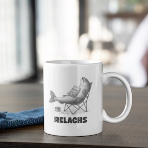 May include: White ceramic mug featuring a humorous illustration of a fish relaxing in a beach chair, holding a bag of chips. The mug has the word "IRELACKS" printed below the image.