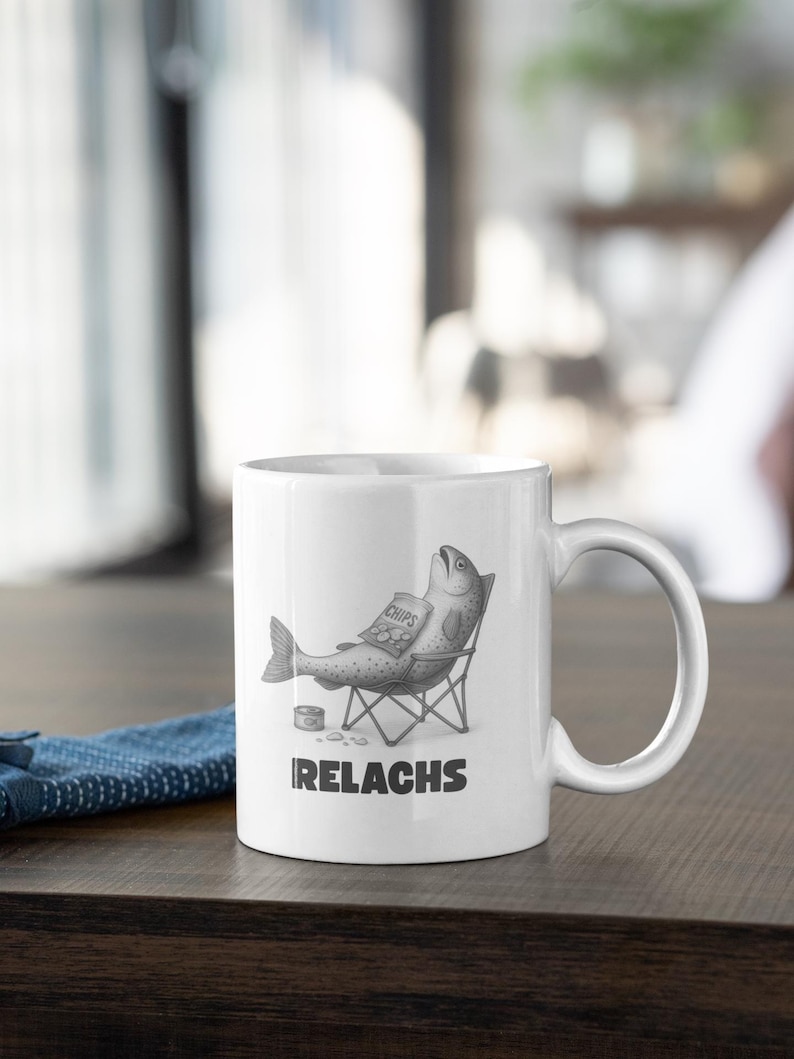 May include: White ceramic mug featuring a humorous illustration of a fish relaxing in a beach chair, holding a bag of chips. The mug has the word "IRELACKS" printed below the image.