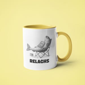 May include: White ceramic mug with a yellow interior and handle. The mug features a black and white illustration of a fish relaxing in a beach chair, holding a bag of chips. The word "RELACHS" is printed below the illustration.