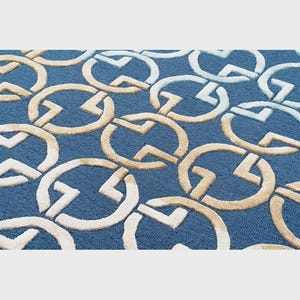 May include: A blue rug with a repeating pattern of interlocking circles and angular shapes in beige and white. The rug has a textured surface and a dense pile, creating a visually interesting and tactile design. The overall aesthetic is modern and geometric.