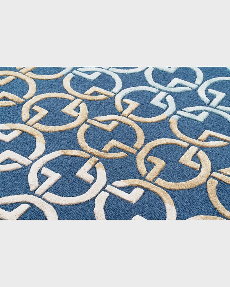 May include: A blue rug with a repeating pattern of interlocking circles and angular shapes in beige and white. The rug has a textured surface and a dense pile, creating a visually interesting and tactile design. The overall aesthetic is modern and geometric.