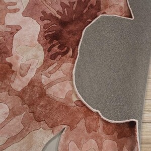 May include: A close-up of a rug with a floral design in shades of pink and brown. The rug's underside is gray, and the edge is visible. The rug is placed on a light brown wooden surface.