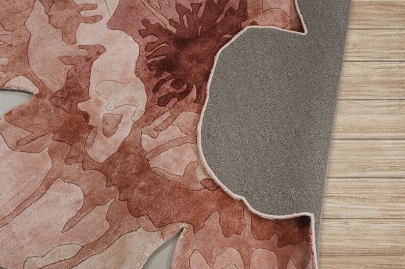 May include: A close-up of a rug with a floral design in shades of pink and brown. The rug's underside is gray, and the edge is visible. The rug is placed on a light brown wooden surface.