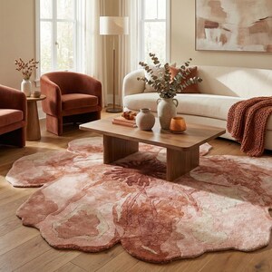 May include: A living room scene featuring a large, floral-shaped rug in shades of pink and cream. Two rust-colored armchairs and a wooden coffee table are arranged around the rug. A white sofa with a rust-colored throw blanket and decorative vases complete the cozy interior.