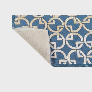 May include: Blue rug with a repeating pattern of interlocking cream-colored circles. The rug's edge is visible, revealing a neutral-colored backing. The design creates a geometric and modern aesthetic, suitable for home decor.