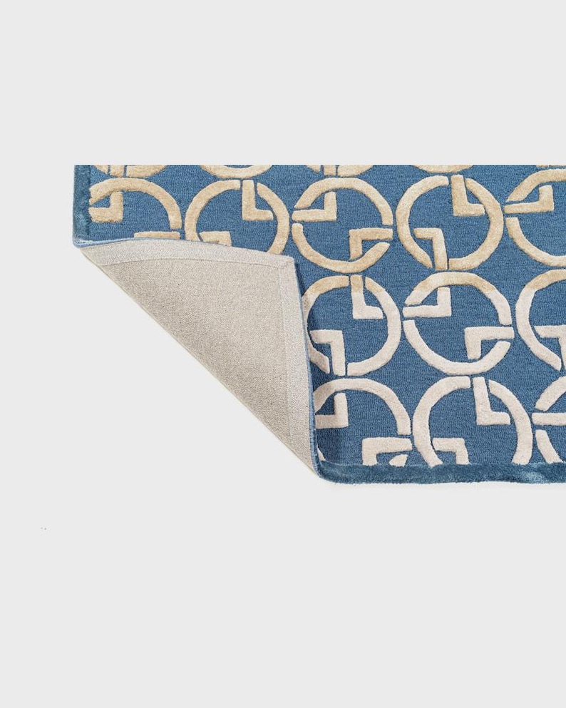 May include: Blue rug with a repeating pattern of interlocking cream-colored circles. The rug's edge is visible, revealing a neutral-colored backing. The design creates a geometric and modern aesthetic, suitable for home decor.