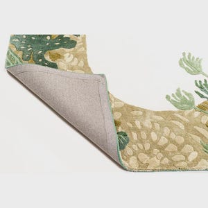 May include: A section of a decorative rug with a botanical design. The rug features a beige background with green and olive-colored floral patterns. The underside of the rug is visible, showing a gray backing and a green border.
