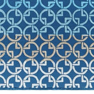 May include: A rectangular blue rug with a repeating pattern of interlocking circles and cross shapes in shades of blue and beige. The rug has a textured surface and a dark blue border. A small green tag is visible on the right side.