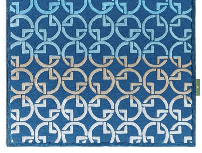 May include: A rectangular blue rug with a repeating pattern of interlocking circles and cross shapes in shades of blue and beige. The rug has a textured surface and a dark blue border. A small green tag is visible on the right side.