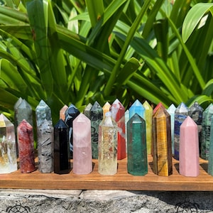 Könnte beinhalten: Eine Sammlung verschiedener Kristallspitzen in unterschiedlichen Farben und Mustern, angeordnet auf einem Holzregal. Die Kristalle umfassen klare, rosa, grüne und gestreifte Varianten, mit einem Hintergrund aus grünem Laub.