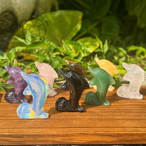 以下が含まれることがあります： 紫、ピンク、黒、緑など、さまざまな色の宝石で彫られたイルカの置物のコレクション。 イルカは、緑の葉の背景を持つ木製の表面に展示されています。
