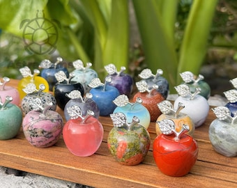 May include: A collection of gemstone apple sculptures in various colors, including red, pink, green, and blue. Each apple has a silver-colored stem and leaf design. The apples are displayed on a wooden surface.