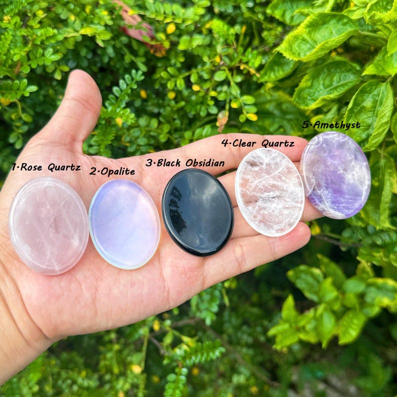 May include: Five polished gemstones: Rose Quartz, Opalite, Black Obsidian, Clear Quartz, and Amethyst. Each stone is oval-shaped and held in a hand against a backdrop of green foliage. The names of the stones are labeled in white text.