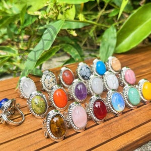 Puede incluir: Una colección de anillos de piedras preciosas con piedras de forma ovalada en varios colores, incluyendo azul, verde, naranja y rosa. Los anillos tienen un engaste plateado y adornado, y se exhiben sobre una superficie de madera.