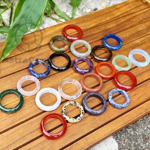 Könnte beinhalten: Eine Sammlung von Edelsteinringen in verschiedenen Farben, darunter Rot, Grün, Blau und Lila. Die Ringe werden auf einer Holzoberfläche präsentiert, die ihre unterschiedlichen Texturen und Muster hervorhebt. Die Ringe bestehen aus verschiedenen Materialien.