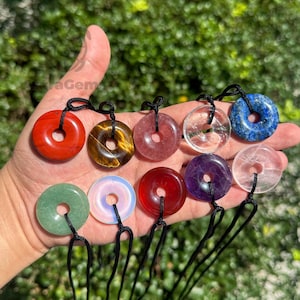 May include: A selection of gemstone donut pendants in various colours, including red, brown, pink, clear, blue, green, and purple. Each pendant is strung on a black cord. The pendants are held in a hand against a blurred green background.
