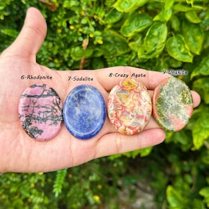 May include: Four polished gemstones: Rhodonite, Sodalite, Crazy Agate, and Unakite. The Rhodonite is pink with black veins, the Sodalite is blue, the Crazy Agate is multi-colored, and the Unakite is green and red. Each stone is oval-shaped.