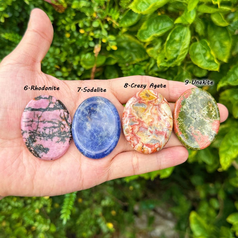 May include: Four polished gemstones: Rhodonite, Sodalite, Crazy Agate, and Unakite. The Rhodonite is pink with black veins, the Sodalite is blue, the Crazy Agate is multi-colored, and the Unakite is green and red. Each stone is oval-shaped.